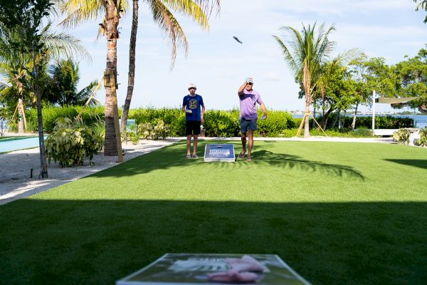 Two people are playing cornhole on a lawn near palm trees and a pool. One is throwing a bean bag towards the target board.