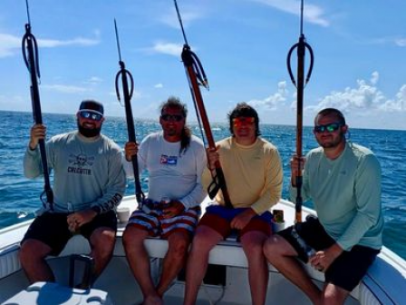 Four people sitting on a boat holding spearguns, with the ocean and a clear sky in the background.