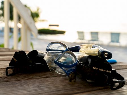 A snorkeling mask and snorkel lie on a wooden deck, with beach chairs and the ocean blurred in the background.