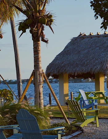 A tropical beach scene with palm trees, a thatched-roof hut, and colorful chairs facing the water, near a calm shoreline.