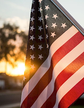 An American flag waving in the breeze at sunset, stars and stripes visible, warm light behind it, patriotic scene.