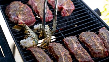 Steaks and lobster tails on a grill, seasoned and being cooked, with tongs handling the meat.