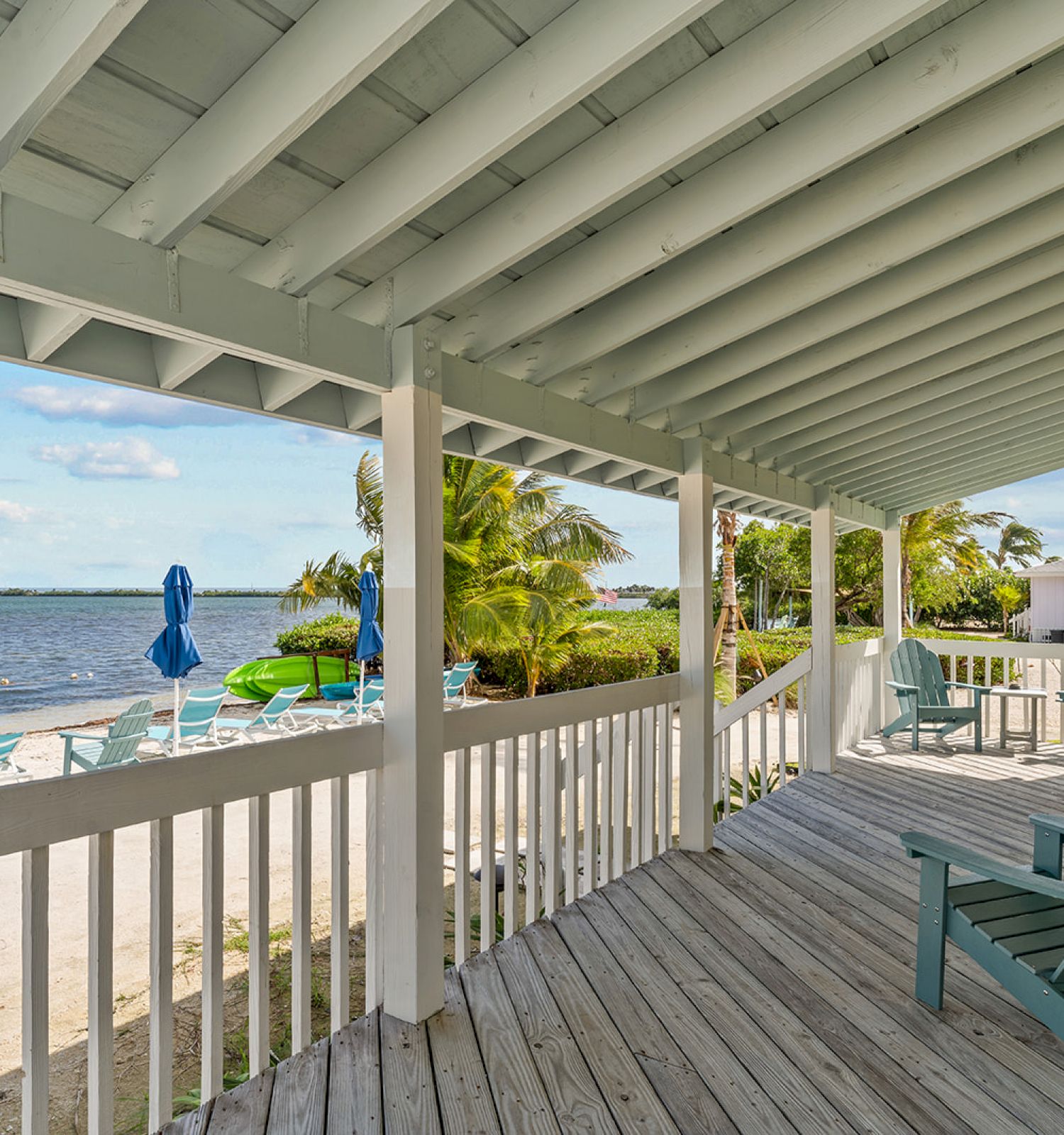 A beachfront porch with blue chairs overlooks a sandy area with more chairs and umbrellas by the water, framed by palm trees and a clear sky.