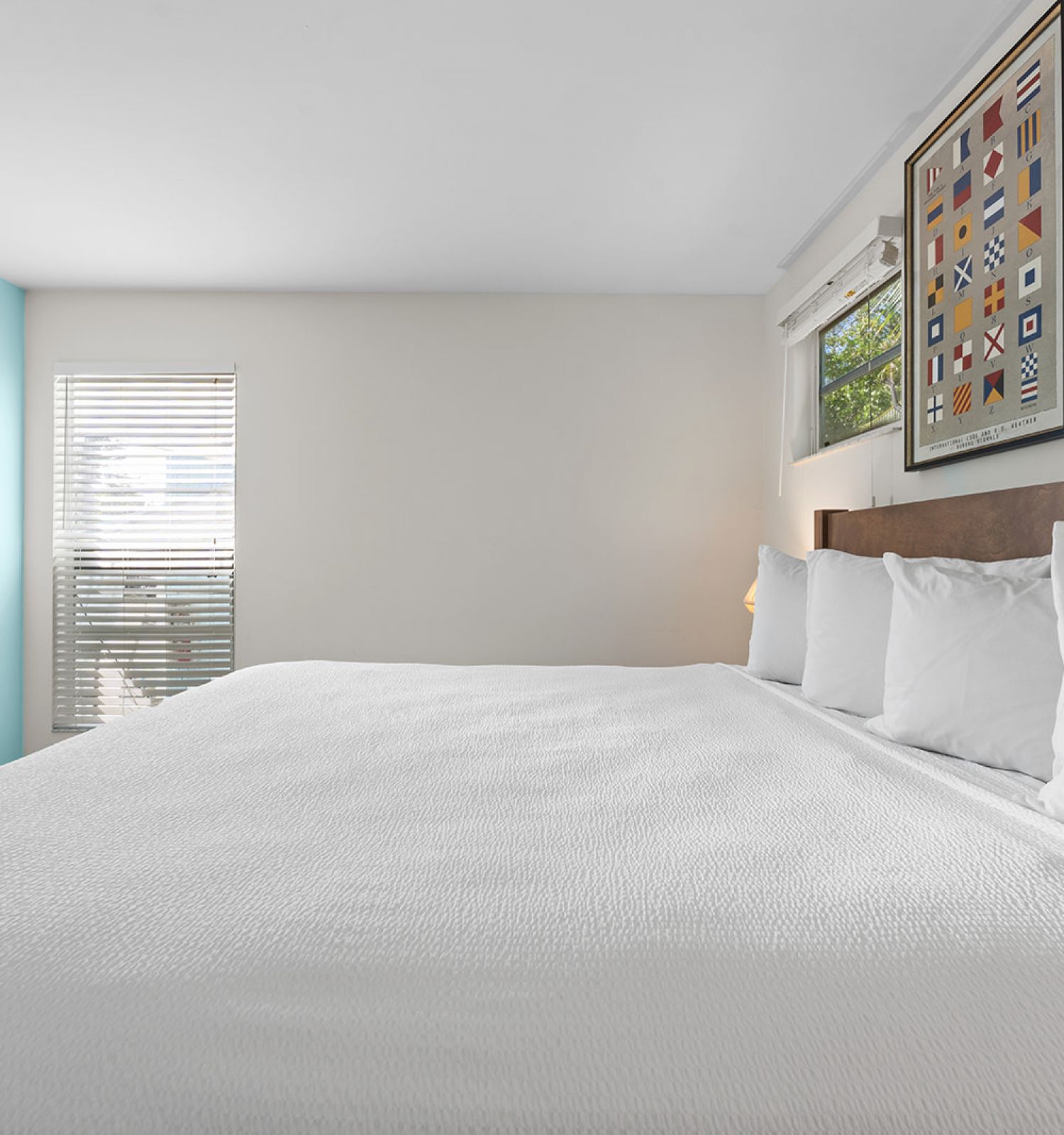 A neatly made bed in a modern bedroom, with artwork above the headboard and sunlight streaming through partially open blinds.