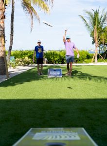 Two people are playing cornhole on a grassy area with palm trees in the background.