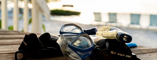 A snorkeling mask and snorkel are placed on a wooden deck by a beach, with lounge chairs and the ocean in the background.