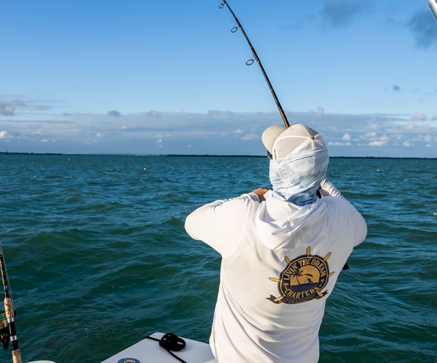 A person is fishing from a boat in the ocean, wearing a hat and long-sleeve shirt under a clear sky.