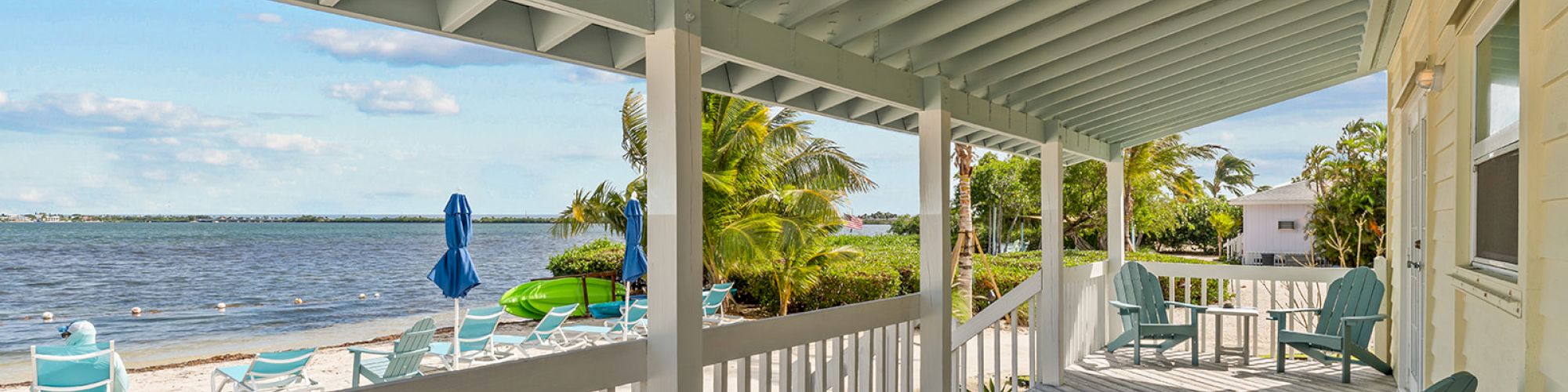 A beachside porch with chairs overlooks sand, water, and distant trees. Blue umbrellas and lounge chairs are on the beach.