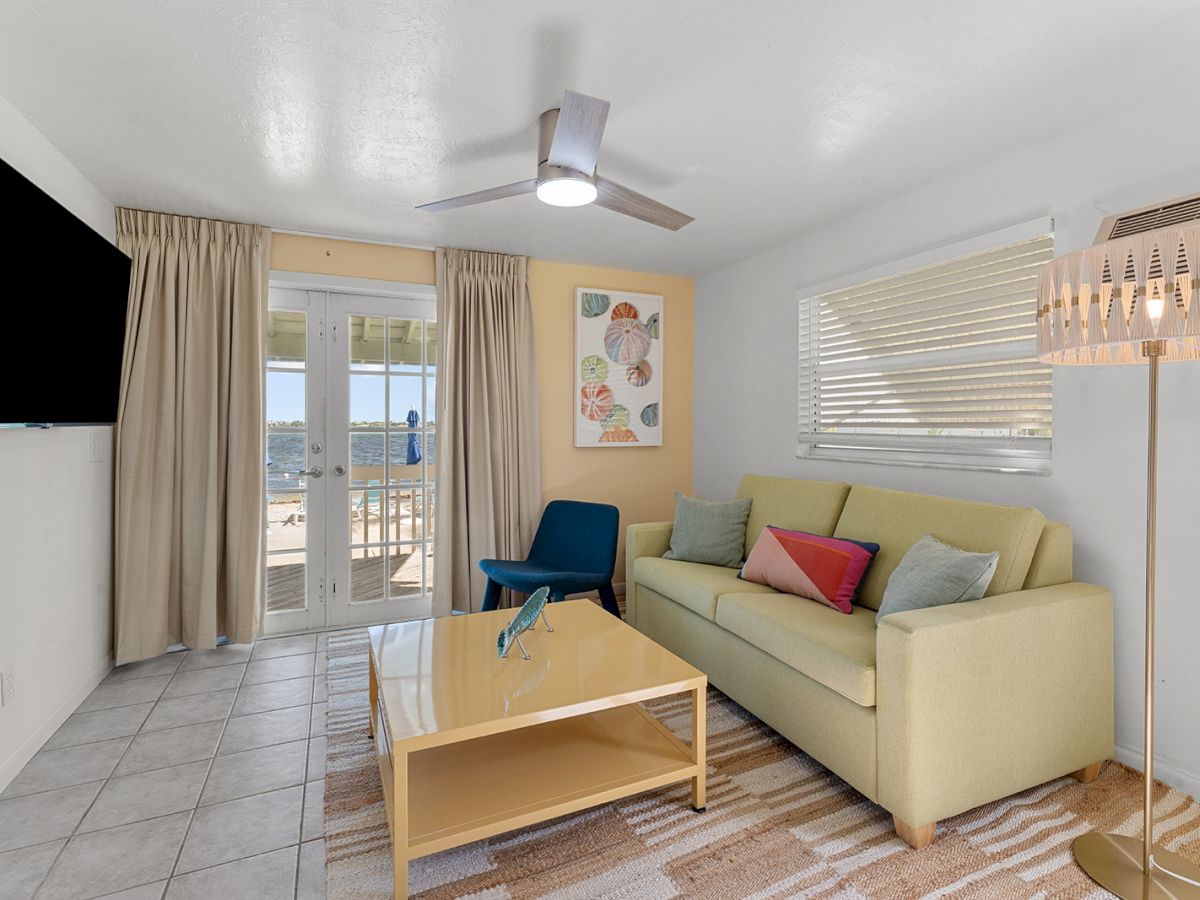 A bright living room with a sofa, coffee table, chair, TV, and balcony access. Wall art and a stylish lamp add decorative touches.