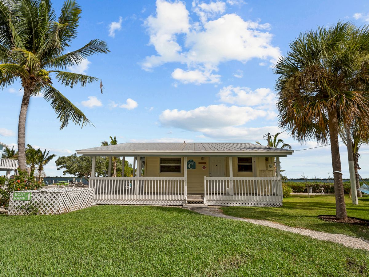 A small house with a porch is surrounded by green grass, palm trees, and a clear blue sky. A body of water is visible in the background.