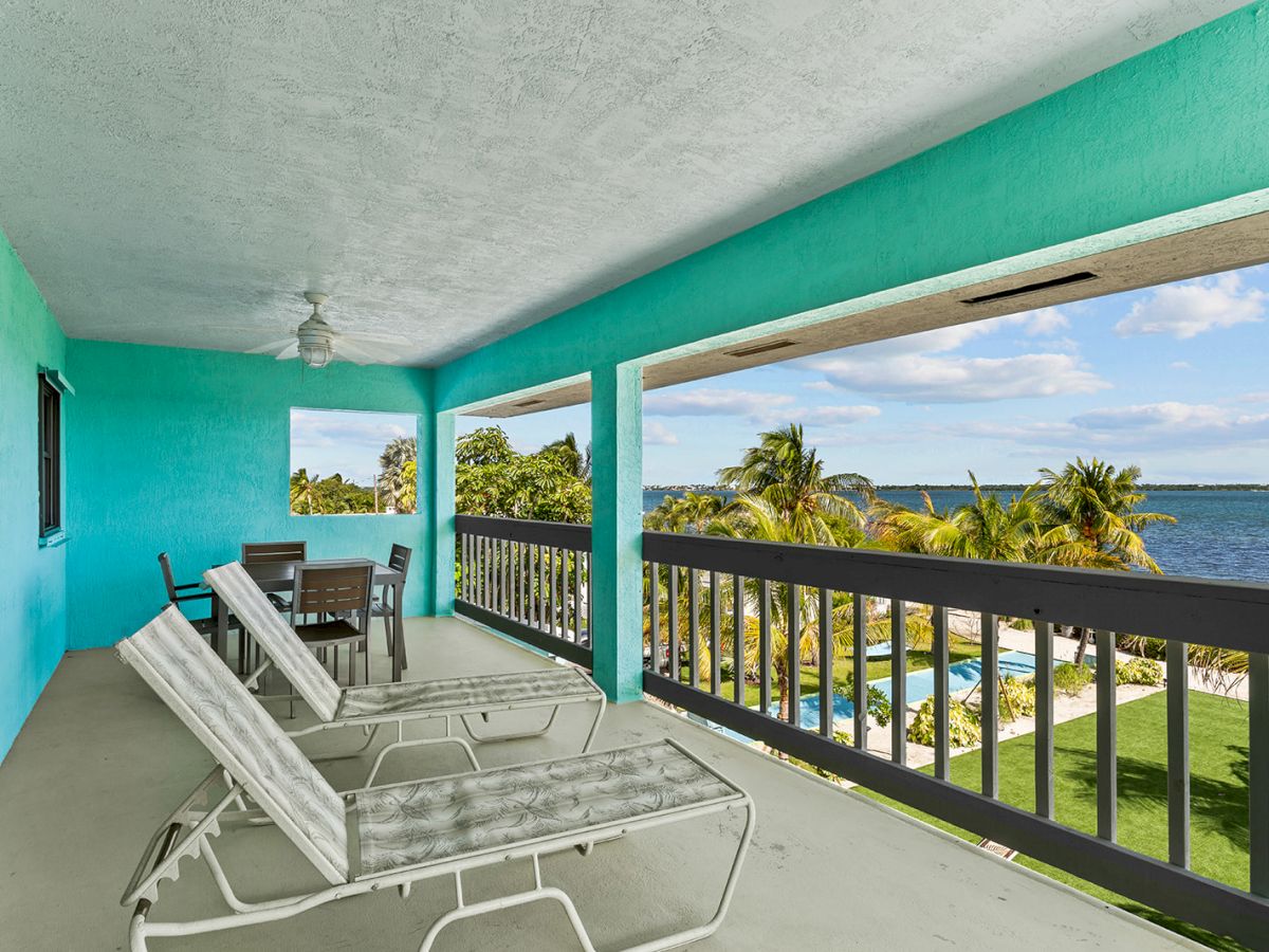 A turquoise-covered balcony with lounge chairs and a table overlooks palm trees, a lawn, and a body of water.