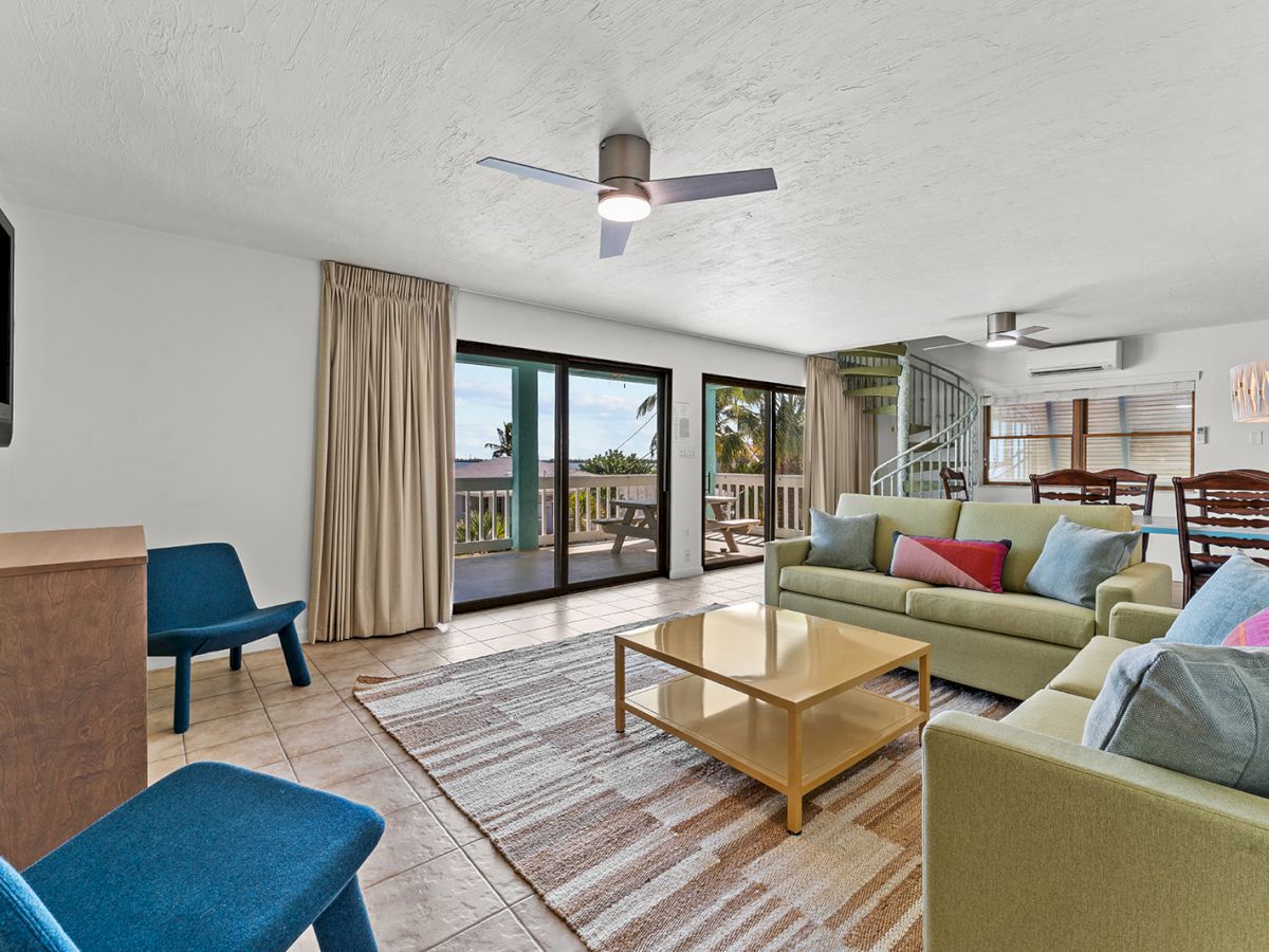 A spacious living room with modern furniture, a TV, and a ceiling fan. Large windows open to a balcony with outdoor views.