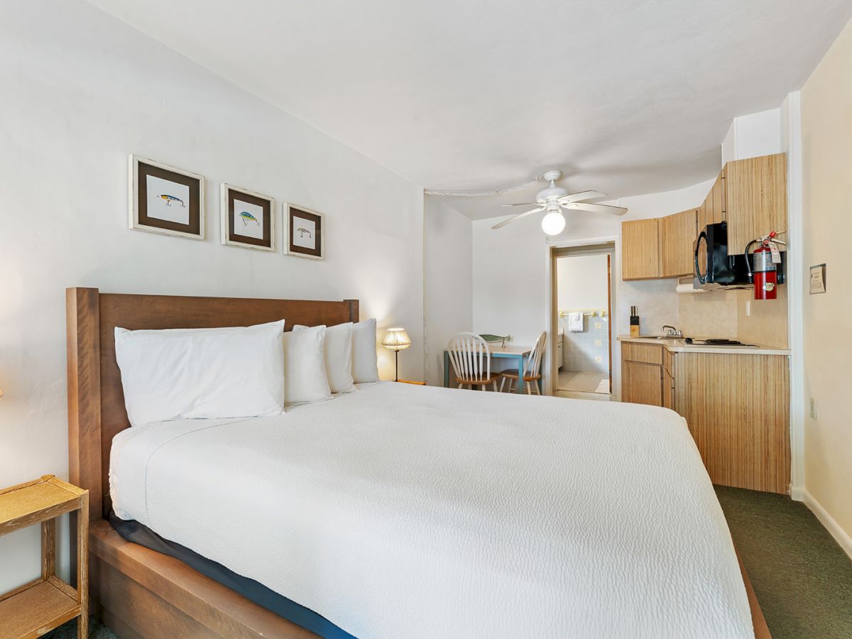 A cozy hotel room with a double bed, wall art, small kitchenette, table with chairs, a ceiling fan, and a mounted TV on the wall.