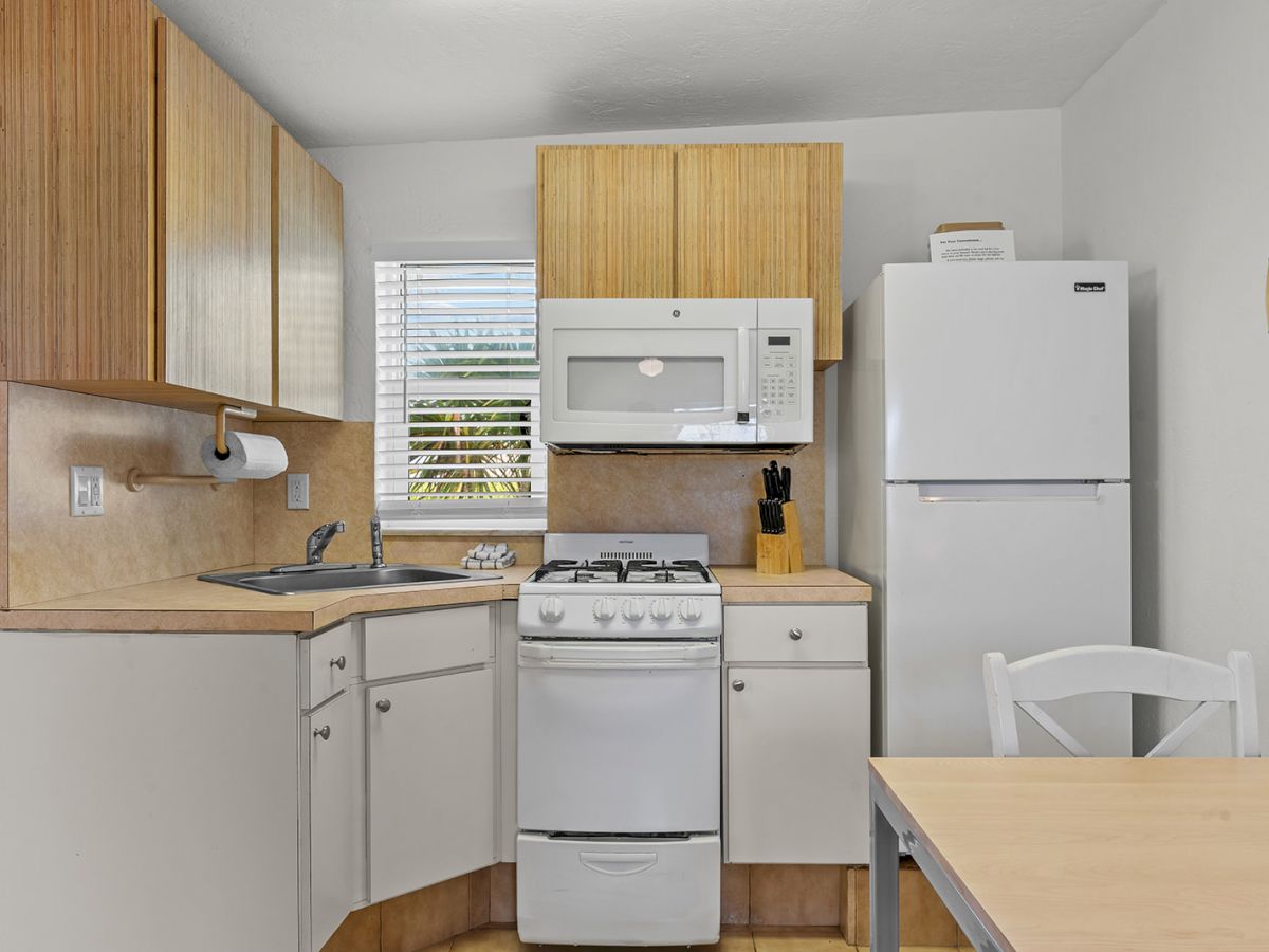 A small kitchen with a stove, microwave, fridge, sink, and cabinets. There’s also a window and a dining table with a chair.