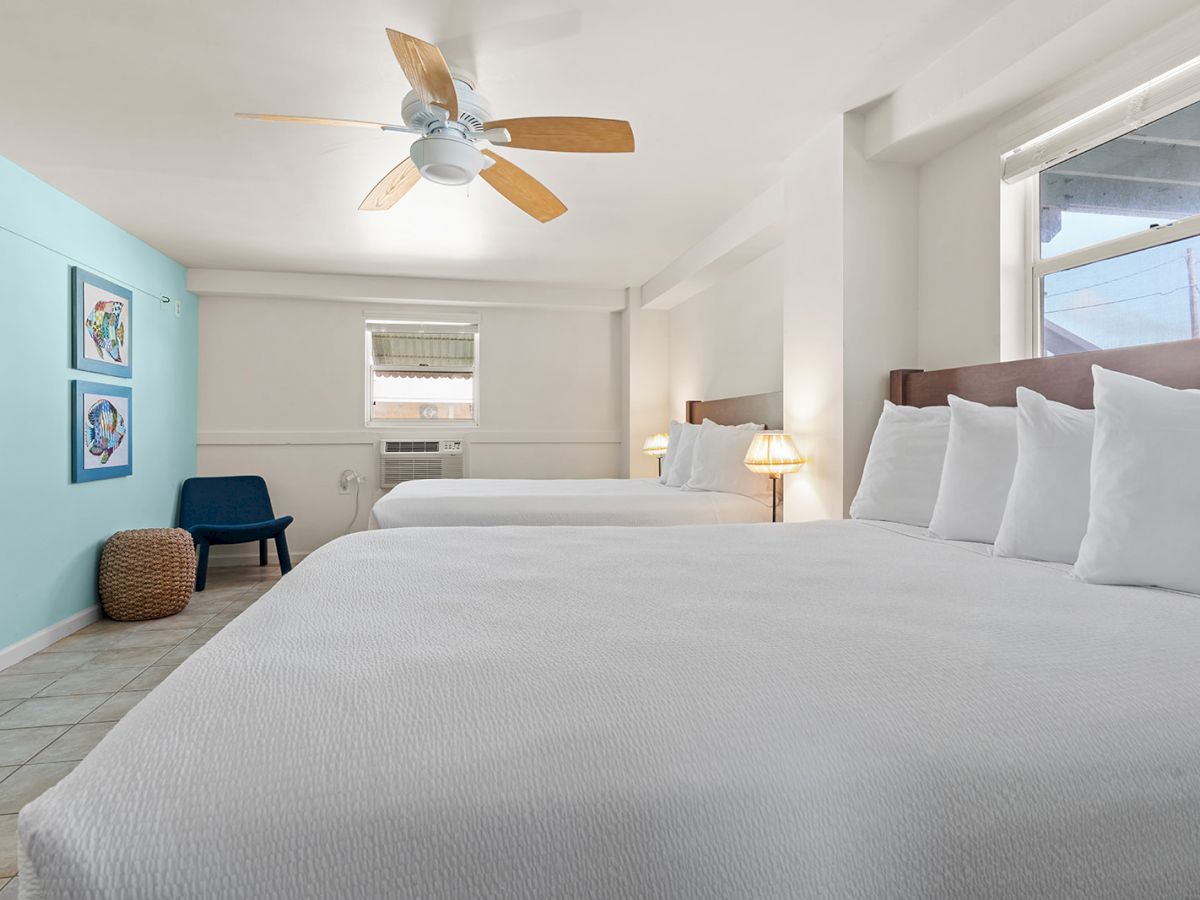 A hotel room with two beds, blue accent wall, ceiling fan, wall-mounted TV, a chair, and windows providing natural light.