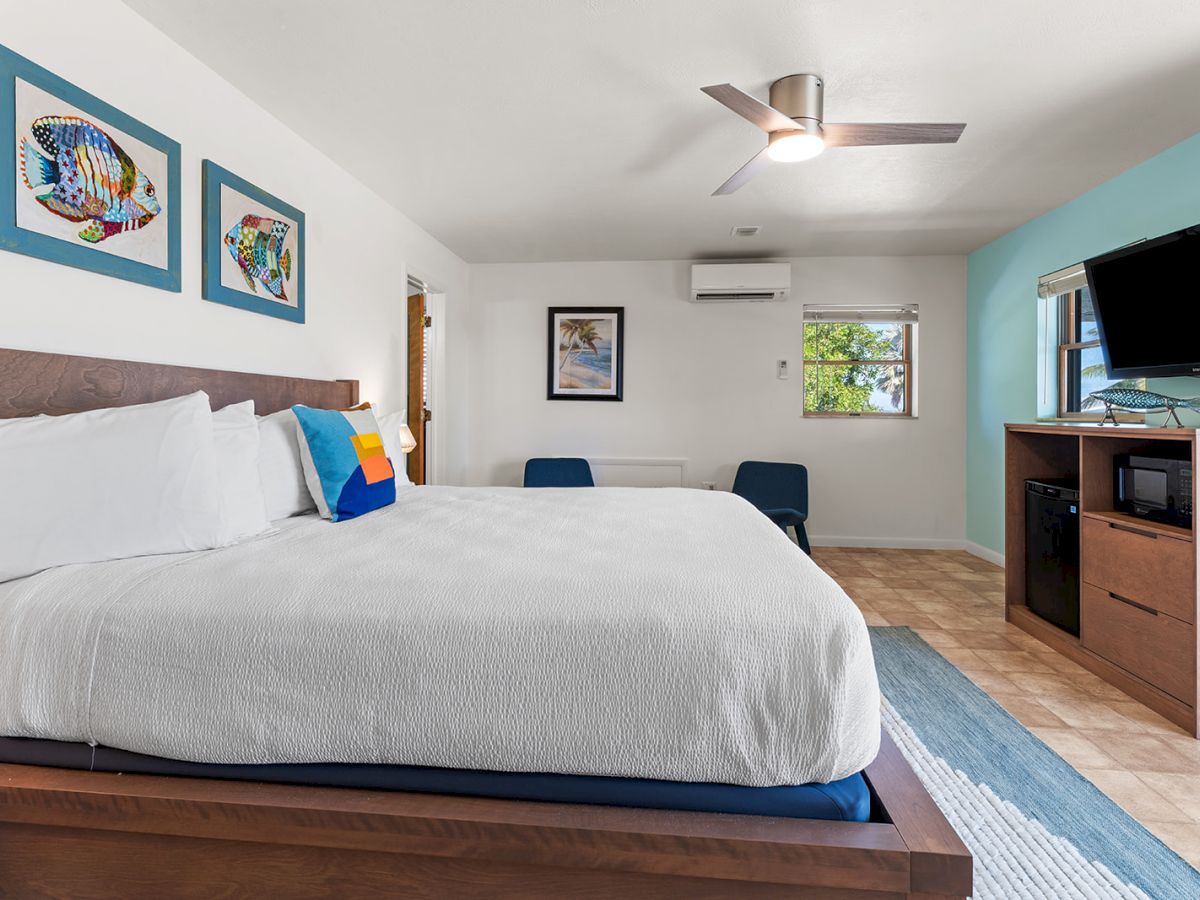 A modern bedroom with a large bed, wall art, TV, microwave, and ceiling fan, featuring light colors and minimalist decor.