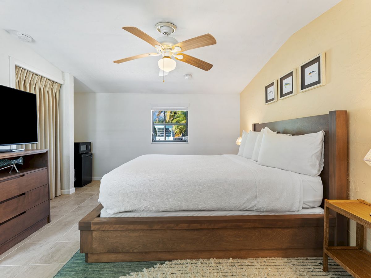 A cozy bedroom with a large bed, ceiling fan, TV, dresser, and small window. Simple decor with framed art and a bedside lamp.