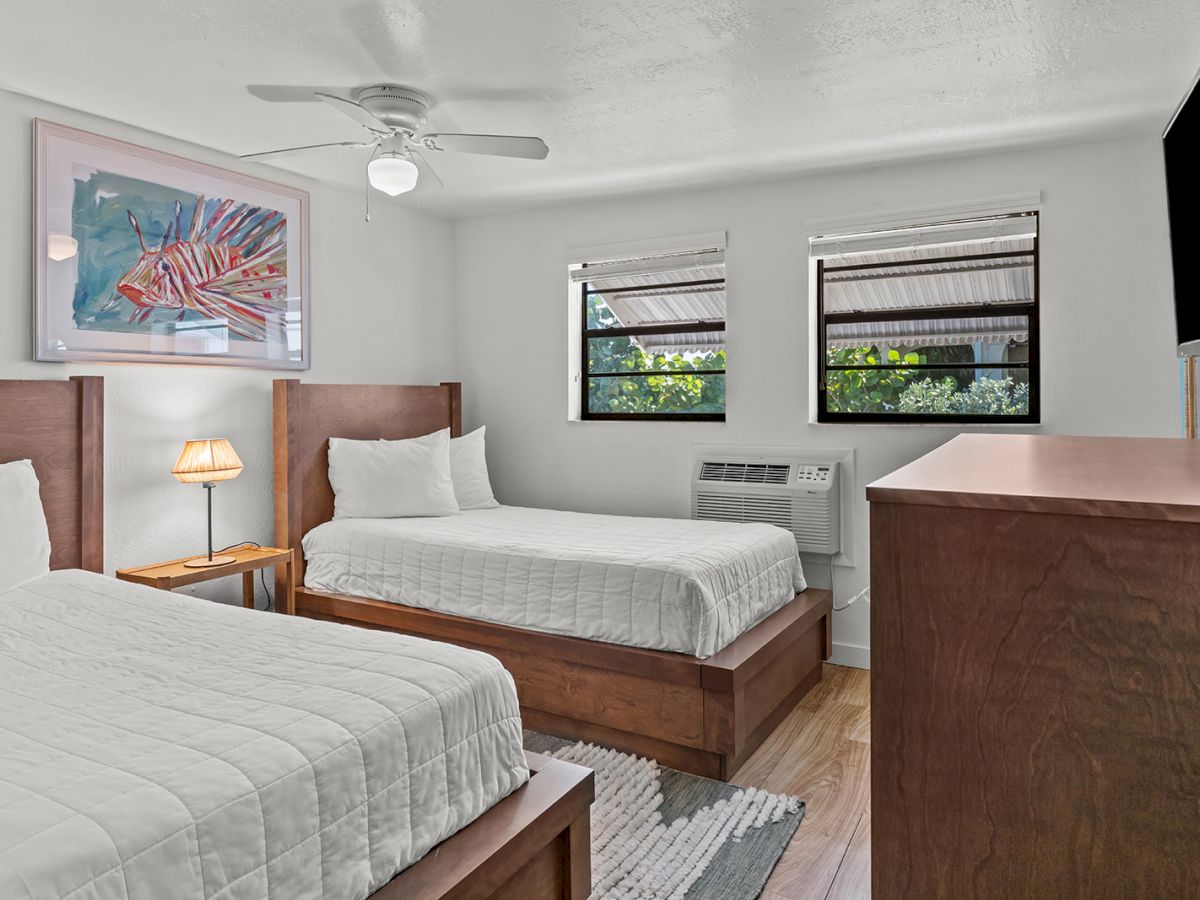 The image shows a bedroom with two beds, a ceiling fan, a nightstand with a lamp, an air conditioner, and a colorful fish artwork on the wall.